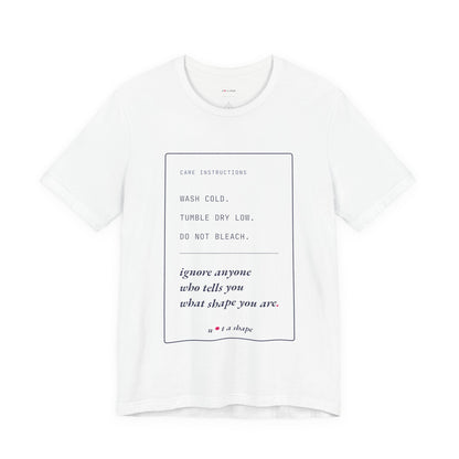 A white short-sleeve crewneck t-shirt with a minimalist graphic print on the front. The print features a thin black rectangular border containing care instructions in black and dark blue text, including 'WASH COLD' and 'ignore anyone who tells you what shape you are.