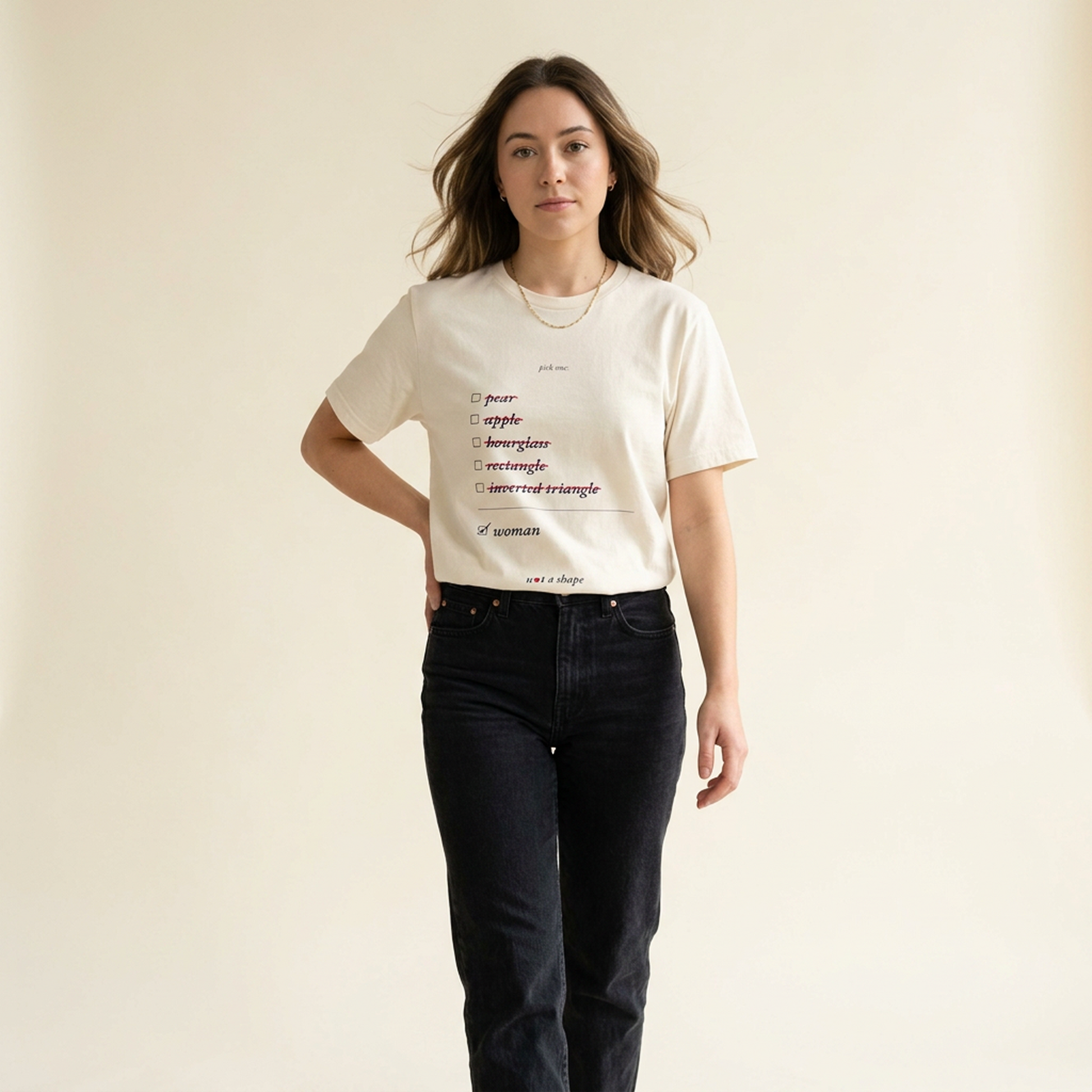 A woman modeling a cream-colored graphic t-shirt with a printed list of adjectives, paired with dark wash jeans, against a solid light beige background.