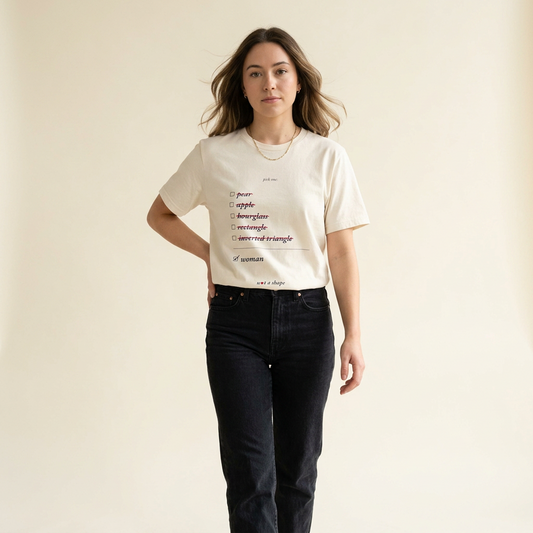 A woman modeling a cream-colored graphic t-shirt with a printed list of adjectives, paired with dark wash jeans, against a solid light beige background.