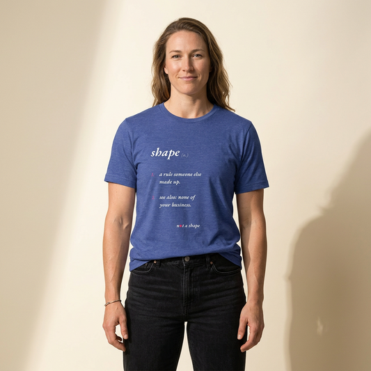 A woman wearing a heathered blue graphic t-shirt with a dictionary-style definition of the word 'shape' printed on the chest, paired with black jeans, standing against a warm beige background with soft shadows.