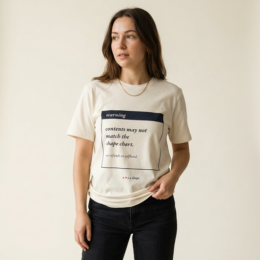 A woman modeling a cream-colored graphic t-shirt with a dark navy and black text print that reads 'warning contents may not match the shape chart. we refund an awkward.