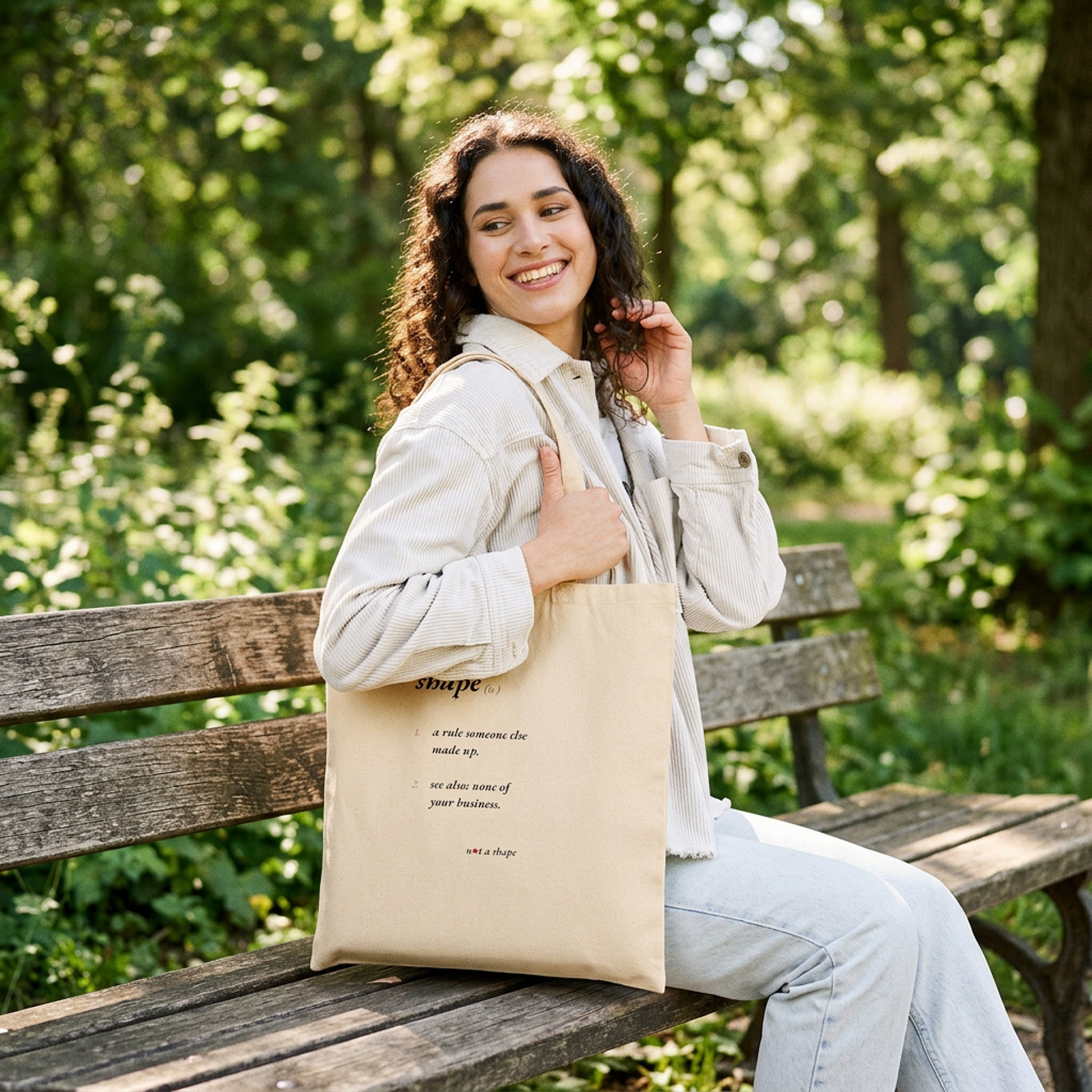 A woman sitting on a wooden park bench holding a beige canvas tote bag with black printed text, set against a blurred green forest background.